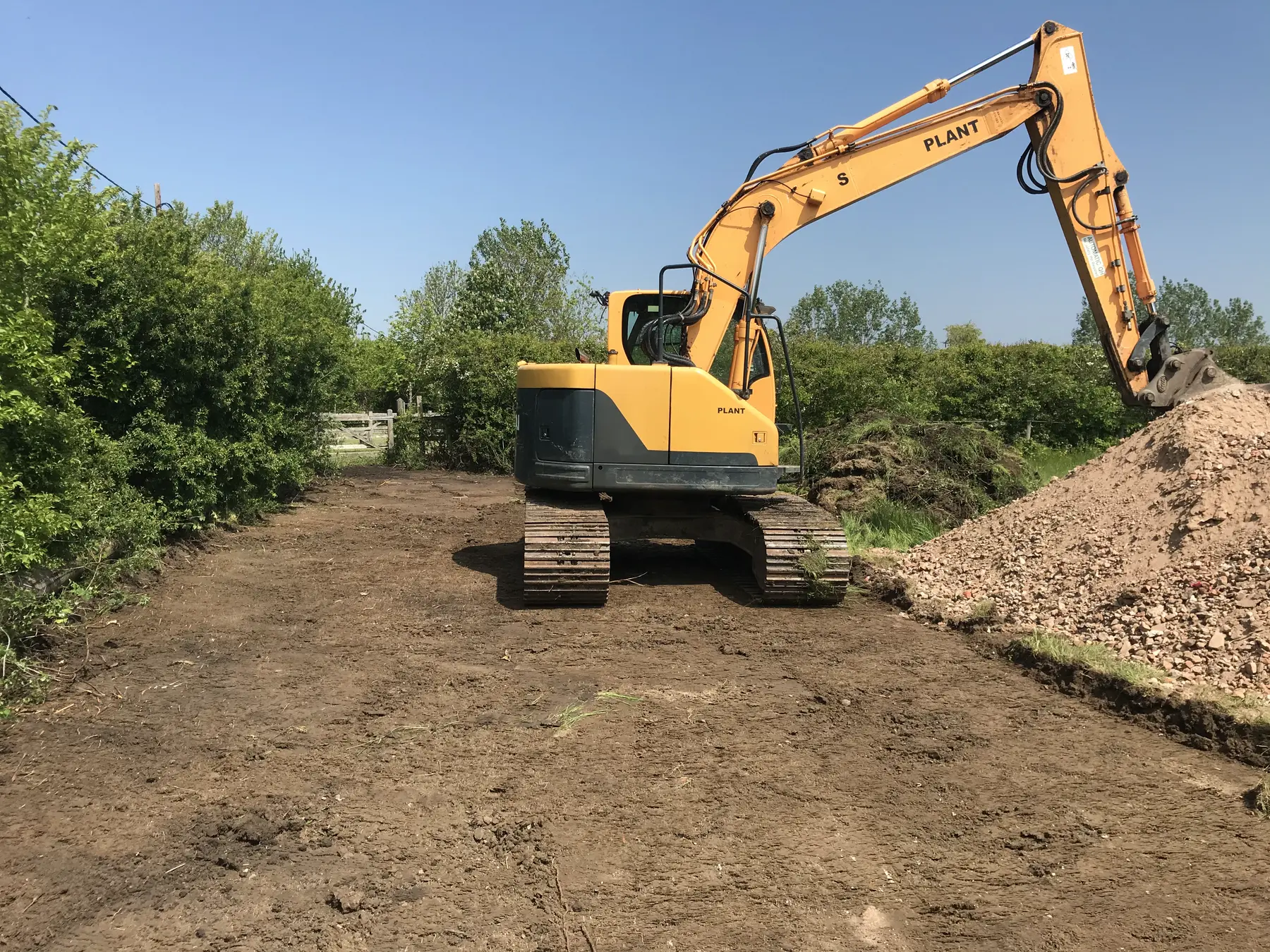 A large yellow excavator on a stripped strip of earth between hedges, next to a heap of hardcore.