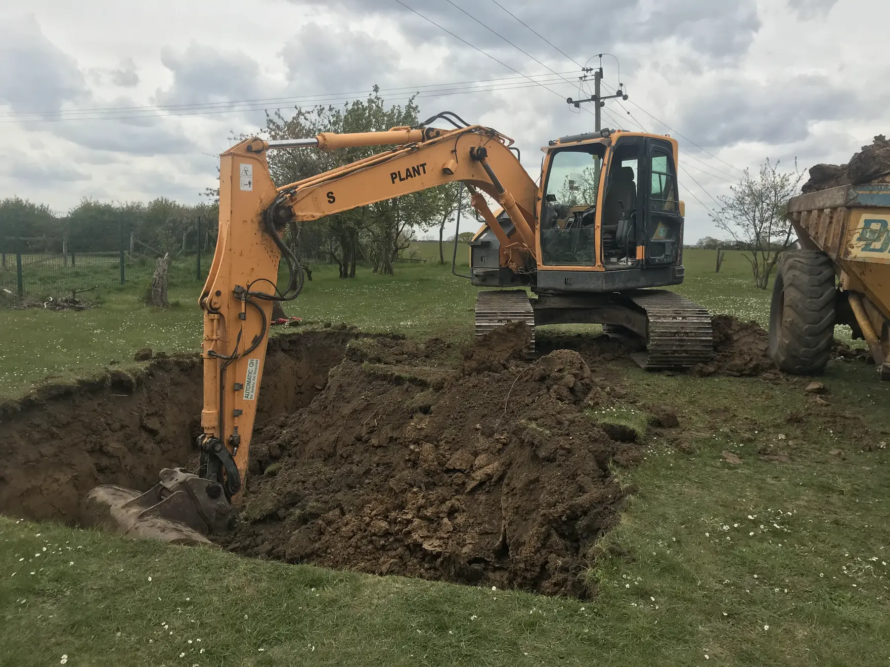 A yellow excavator digs a wide, deep hole in a grass paddock beside a dumper.