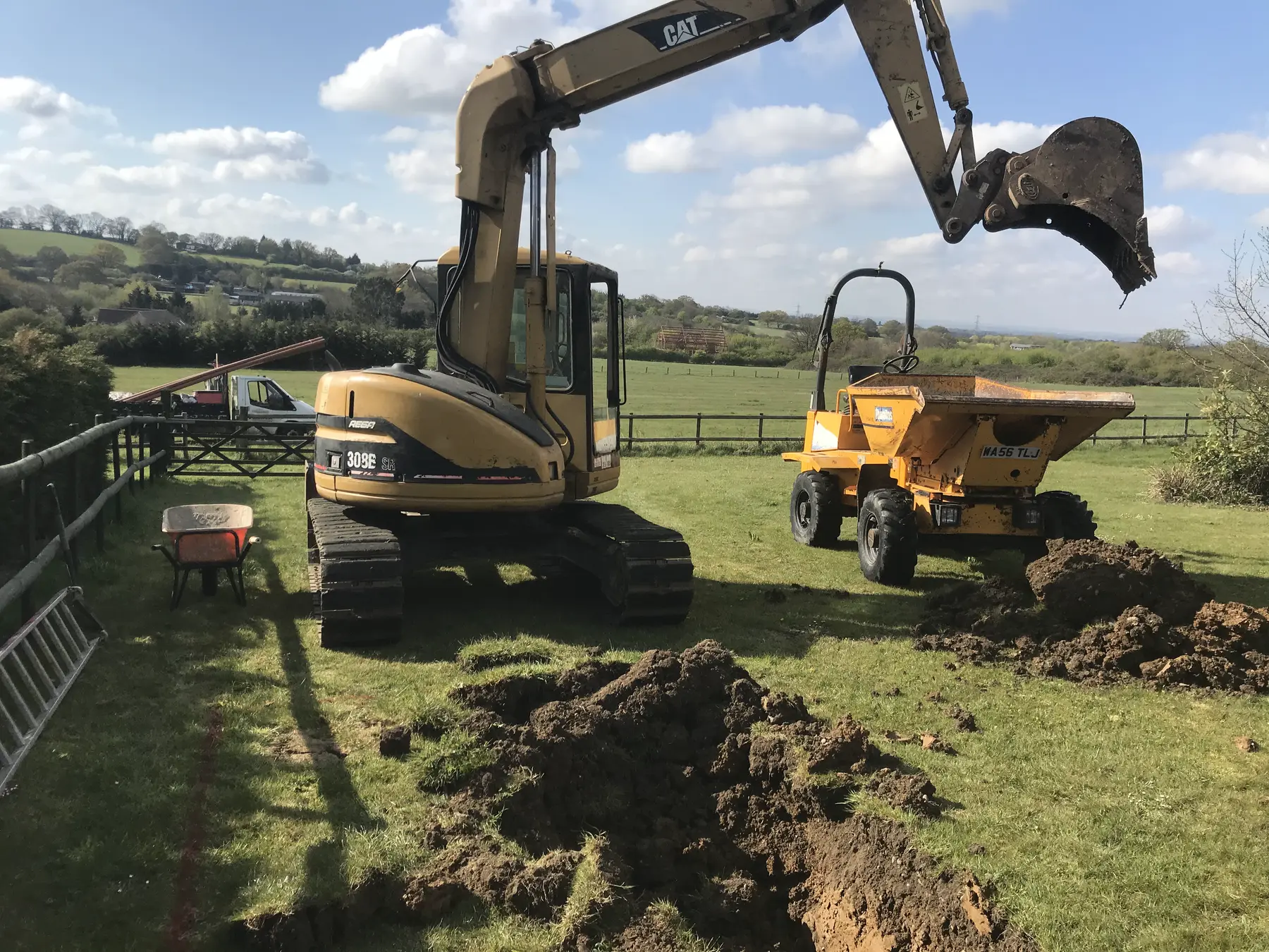 A yellow tracked excavator with its bucket raised over fresh spoil in a fenced paddock, a dumper parked behind.