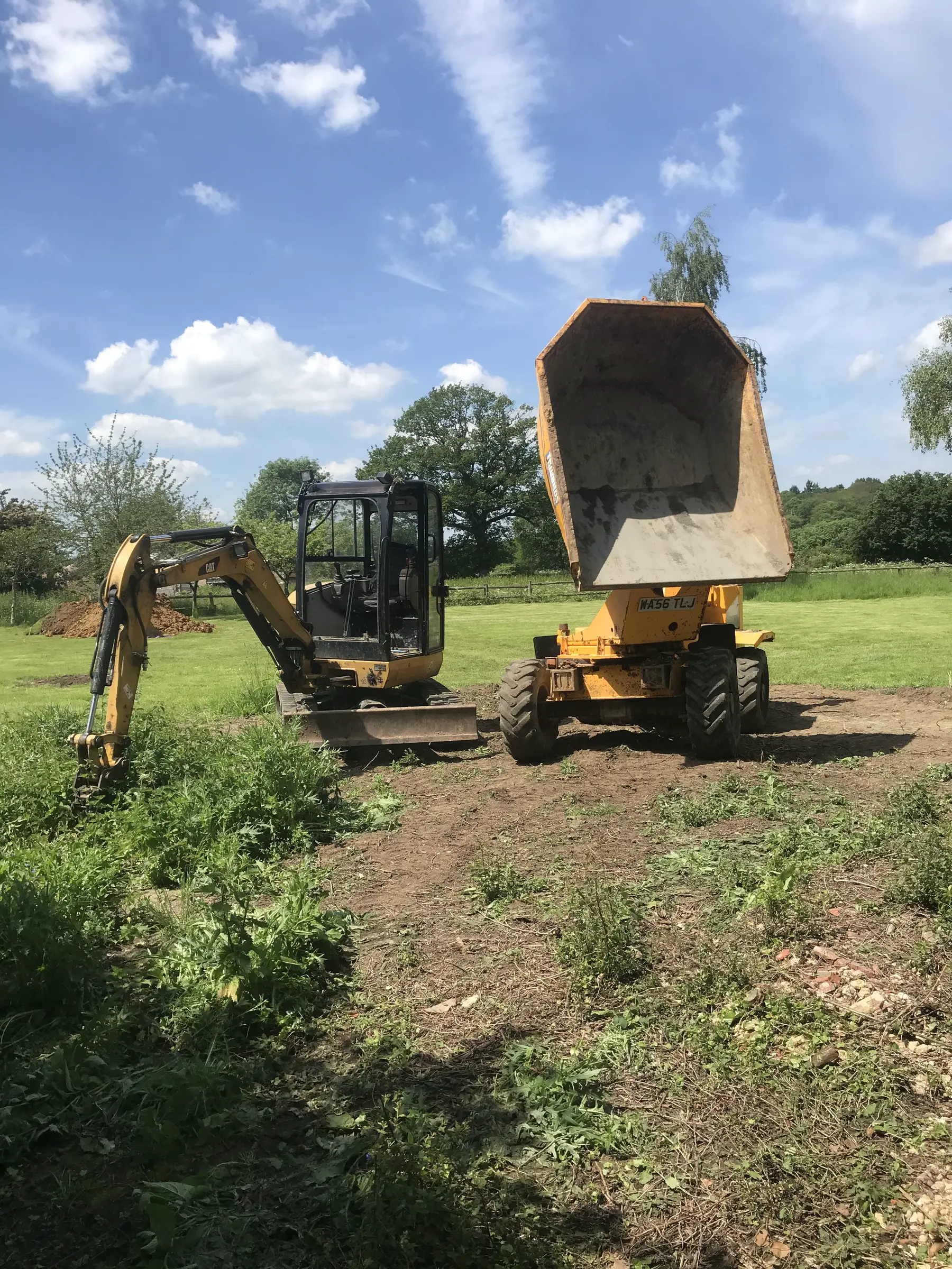 A dumper with its skip tipped high beside a mini excavator in a mown field under a blue sky.