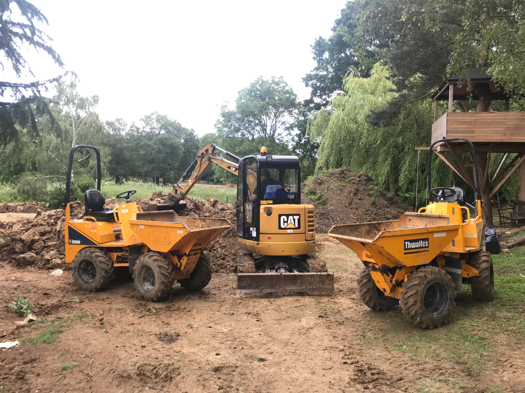 A mini excavator between two yellow dumpers at a garden dig beside a tree house.