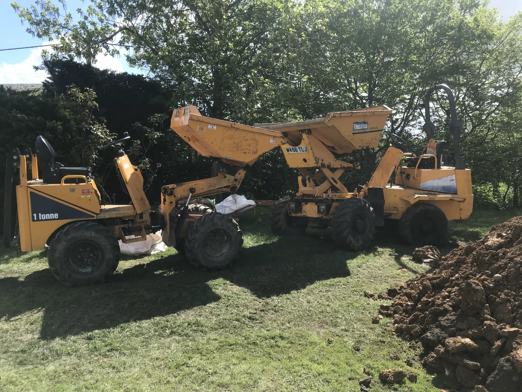 Three yellow site dumpers parked side by side on grass next to a heap of clay.