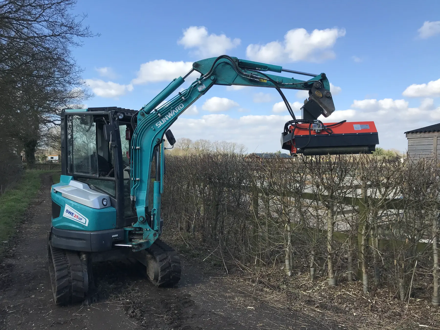 A teal mini excavator trimming a hedge with a flail head on its arm.