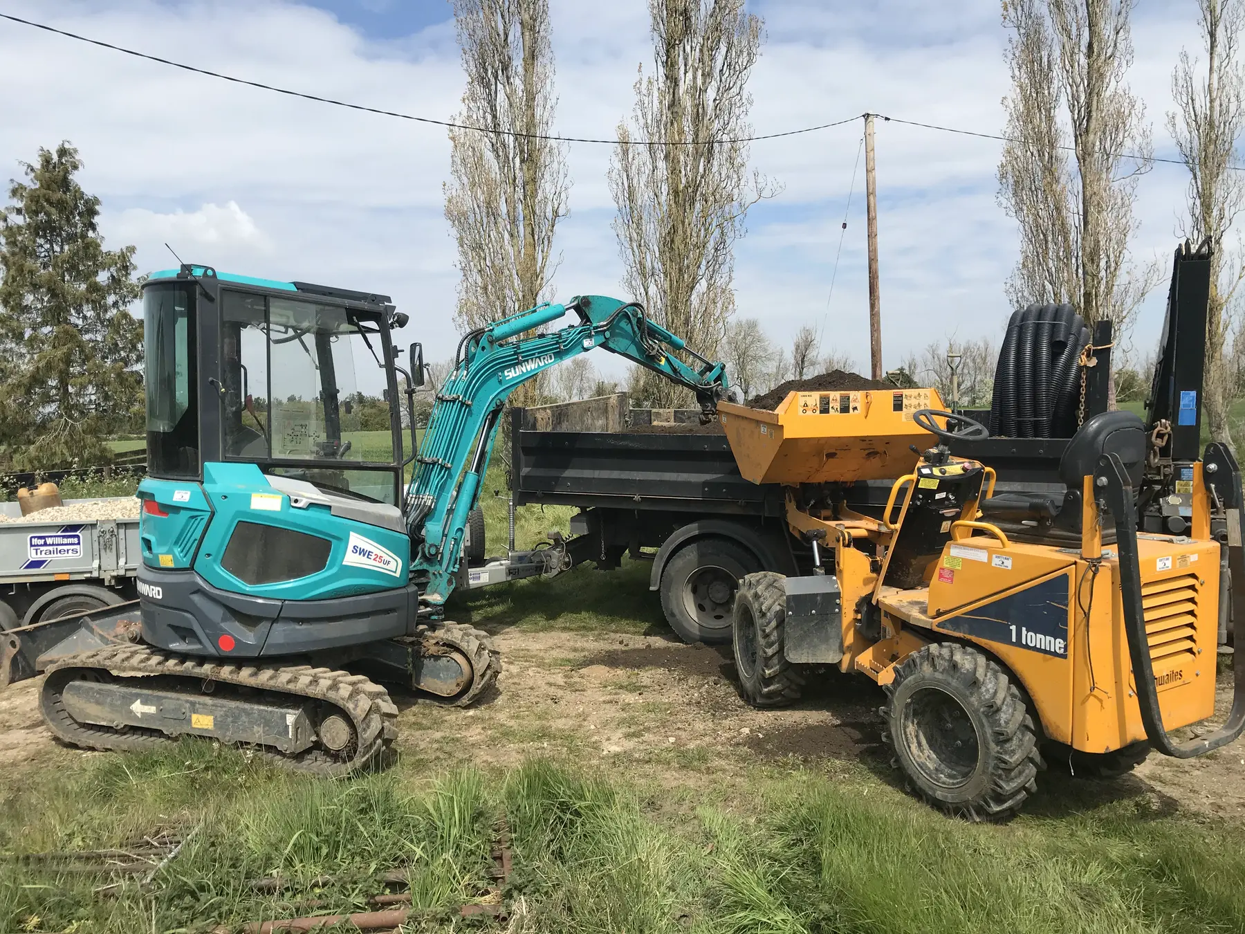 A teal mini excavator loads soil into a raised one-tonne dumper beside a tipper truck.