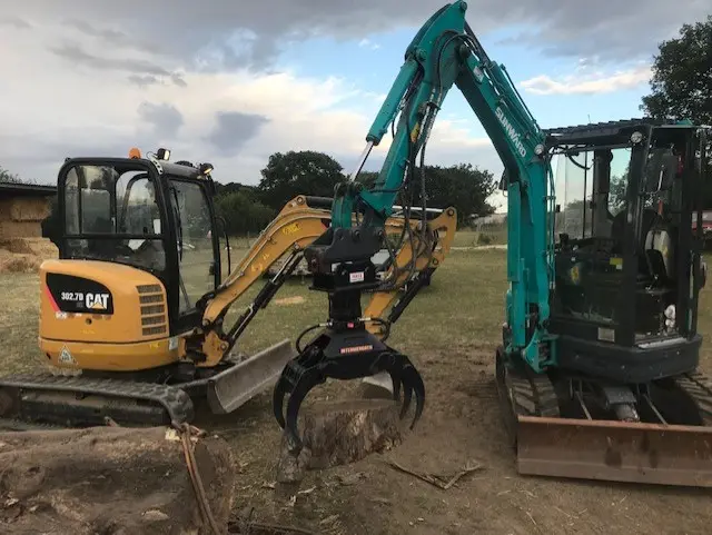 A mini excavator with a hydraulic grab holding a tree stump, a yellow mini digger beside it.