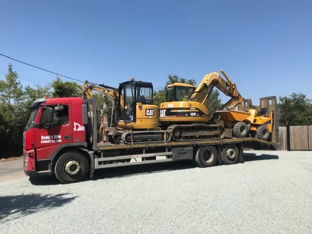 A red beavertail lorry lettered Scott’s Hire carrying two yellow excavators and a dumper.