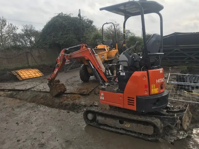 An orange micro excavator digging in a wet yard, a dumper behind.