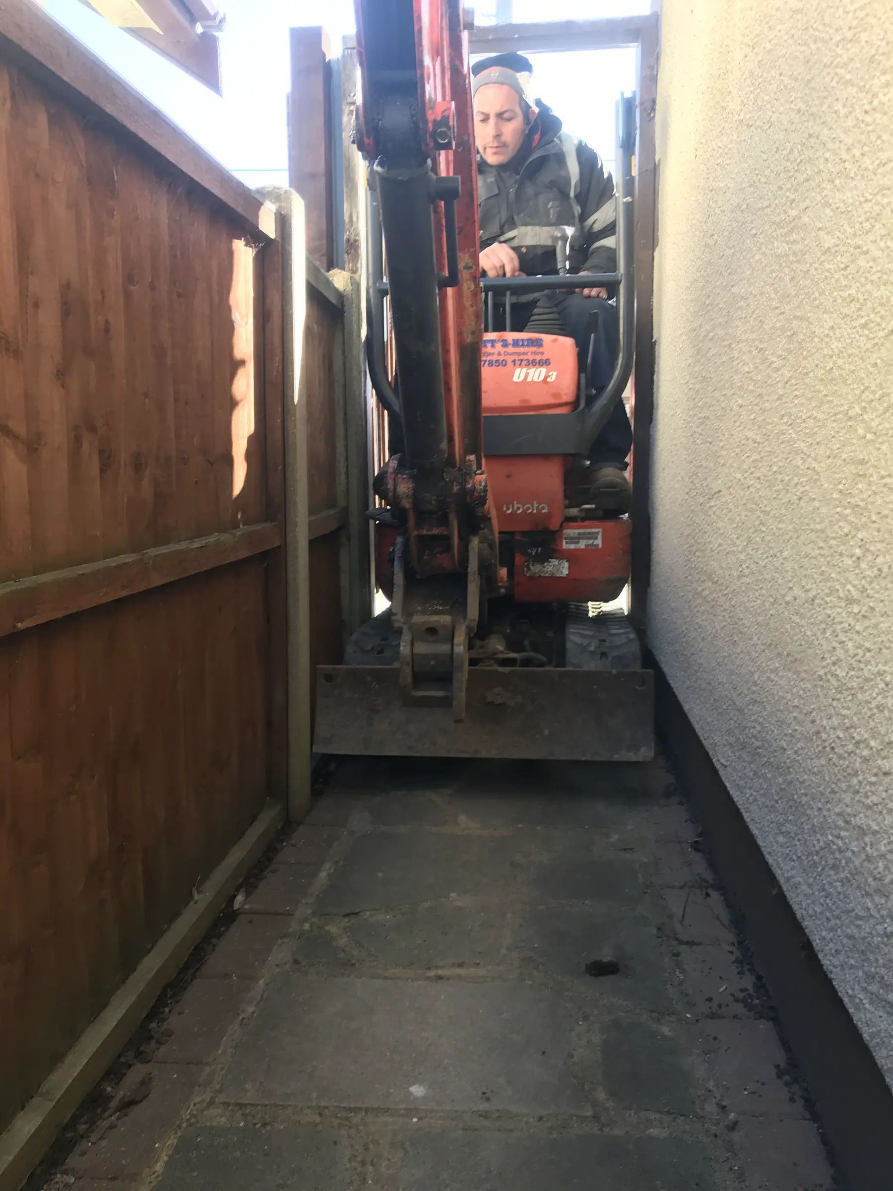 An operator drives a small orange micro excavator down a narrow paved passage between a wooden fence and a house wall.
