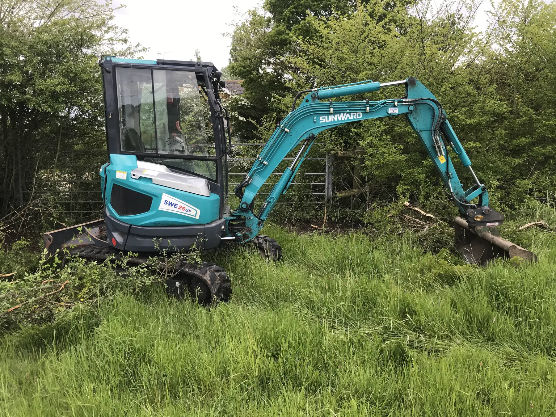 A teal mini excavator with a cab working in long grass by a field gate.