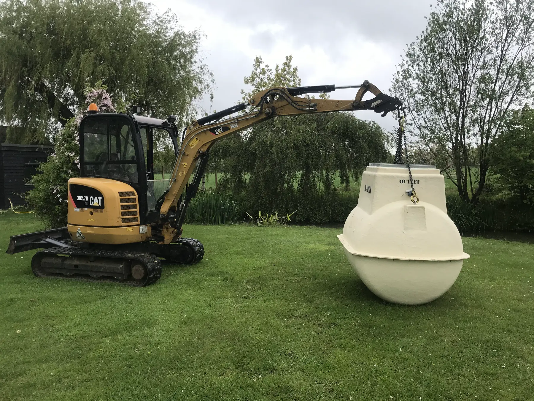 A mini excavator lifts a cream sewage treatment plant tank on a chain across a lawn.