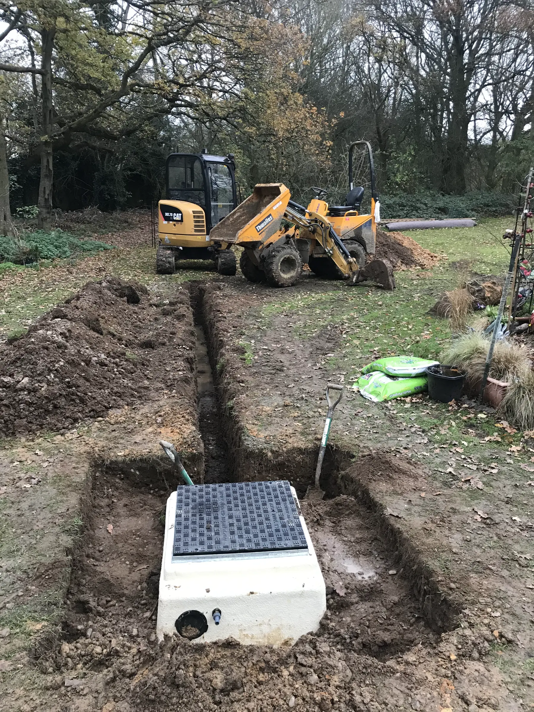 The top of a sewage treatment plant set in a fresh trench, with a mini digger and dumper behind under autumn trees.